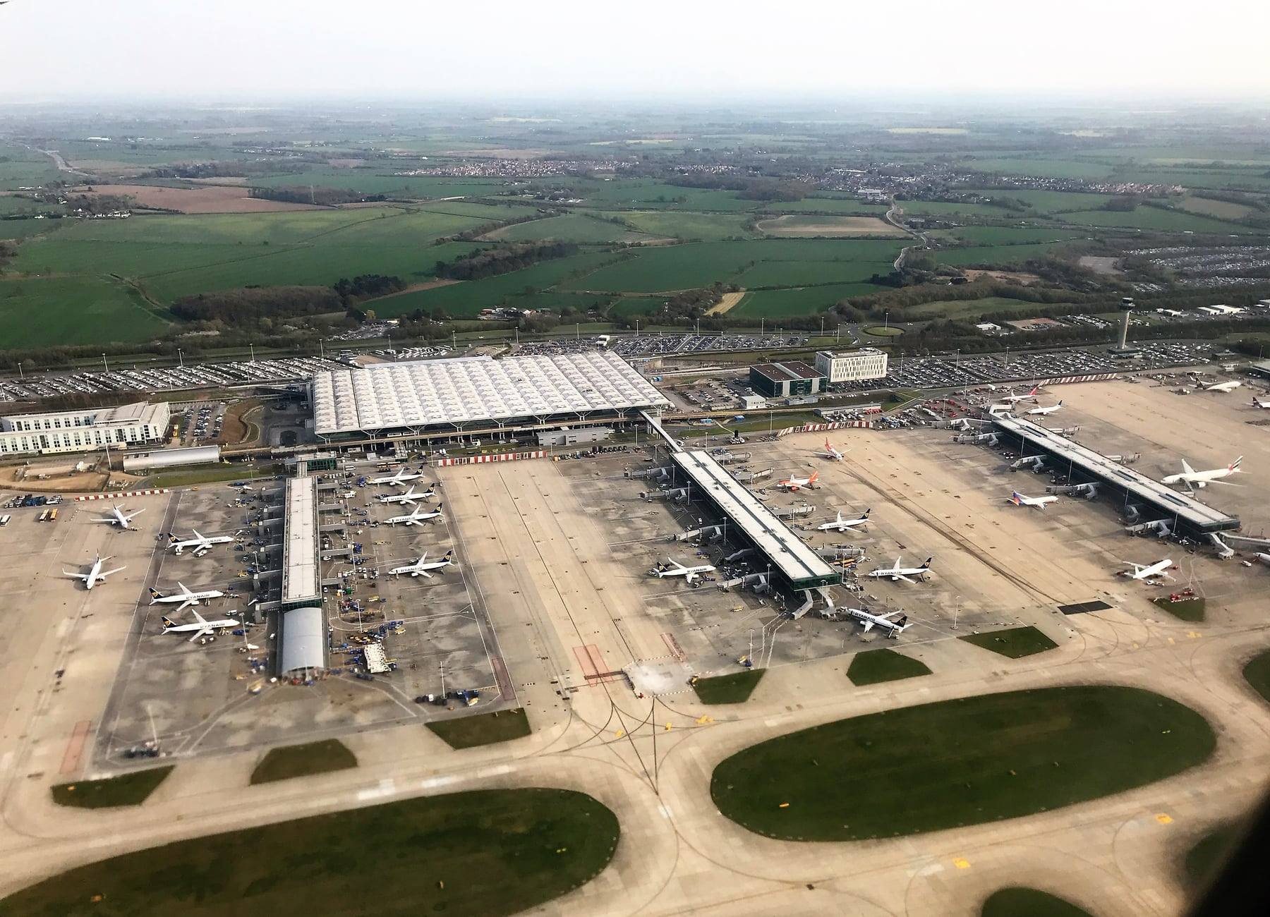 An aerial view of London Stansted Airport, showing the terminal, piers and runway